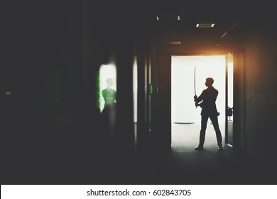 Silhouette Of Boss Or Businessman In Formal Suit Standing In Dark Office Corridor Interior With Reflections And Green Lights And Holding Katana Sword Like Japanese Samurai Warrior Ready To Fight