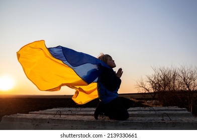 silhouette of boy on his knees, hands folded in prayer, appeal to God. Behind child's back, Ukrainian flag flutters in wind. Children against war. Ukrainian child asks for peace for his homeland