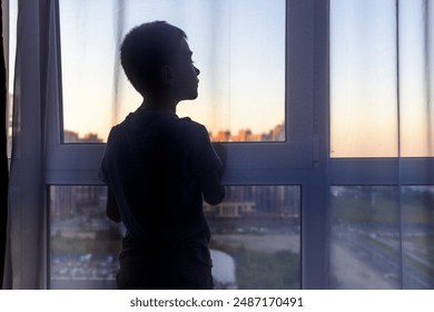 Silhouette of boy standing at window at dawn looking at something interesting. City life. - Powered by Shutterstock