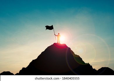 Silhouette Of Businessman Holding Flag On The Top Of Mountain With Over Blue Sky And Sunlight. It Is Symbol Of Leadership Successful Achievement With Goal And Objective Target.