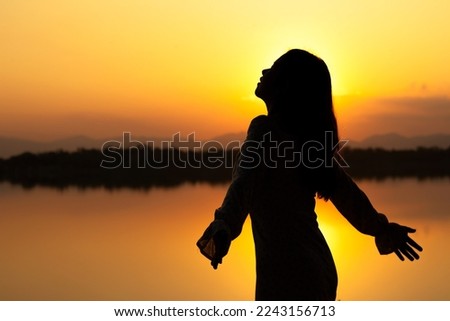 SILHOUETTE: Calm beautiful smiling young asian woman in white dress breathing fresh air outdoor on peaceful evening. relaxing with eyes closed, feeling alive, breathing, dreaming.
