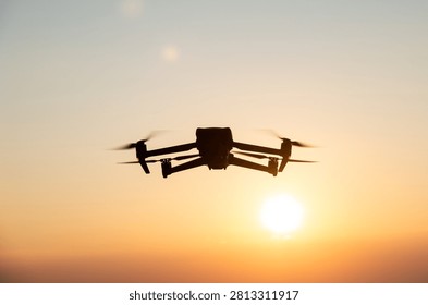 Silhouette of a camera drone hovering in flight against a warm golden sunset sky with the glowing sun and motion-blurred propellers.
