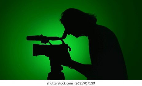 A silhouette of a camera operator looking through a professional camera against a vibrant green background.