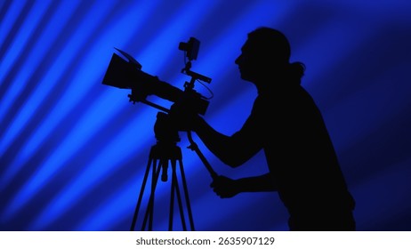 A silhouette of a camera operator working behind a professional camera against a dynamic blue background.