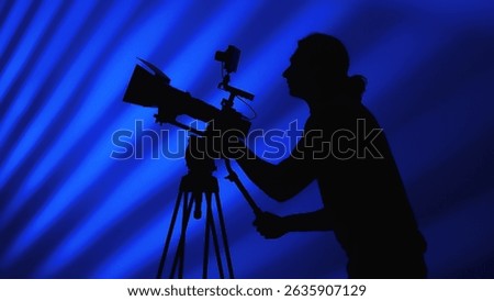 A silhouette of a camera operator working behind a professional camera against a dynamic blue background.