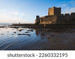 Silhouette of Carrickfergus Castle, Northern Ireland, wide angle lens with sunrise, blue sky, water reflections and horizon with a woman walker hiker in winter red jacket and hat