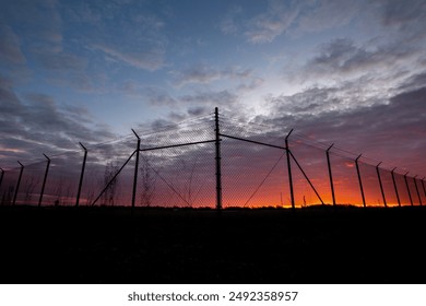 Silhouette of chain link fence with barbed wire at sunset - Powered by Shutterstock