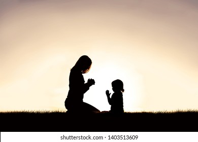 A silhouette of a Christian mother teaching her young child to pray as they sit peacefully outside, against the sunset in the sky.