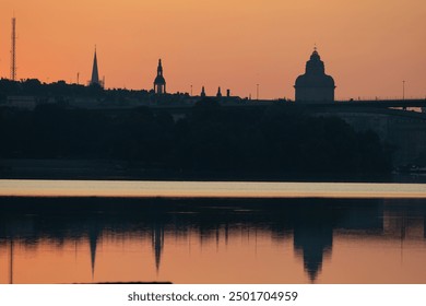 Silhouette of a city skyline at sunset, featuring various church spires and buildings against a warm orange sky. The river reflects the colors, creating a serene atmosphere. - Powered by Shutterstock