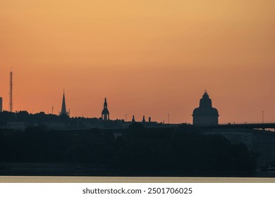 Silhouette of a city skyline at sunset, featuring various church spires and buildings against a warm orange sky. The river reflects the colors, creating a serene atmosphere. - Powered by Shutterstock