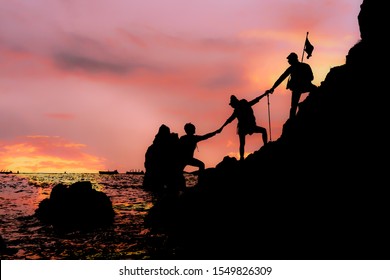 Silhouette Of Climbing Team Helping Each By Hand Pull While Climb Mountain Rock Cliff In A Sunset. Teamwork And Help Concept.