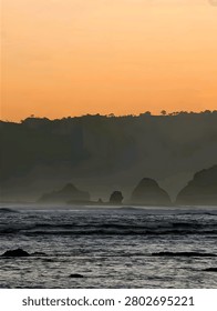 Silhouette of coastal cliffs and sea stack rock formations in misty ocean water under an orange sunset sky