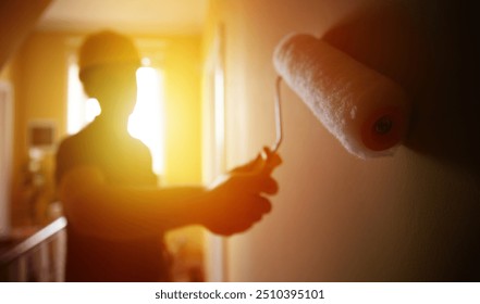 A silhouette of a construction worker painting a wall with a roller is backlit by warm, golden sunlight streaming through a window. The focus on the roller creates a dramatic and artistic effect. - Powered by Shutterstock