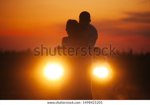 Silhouette of couple hugging in a field, on car lights\
in evening 