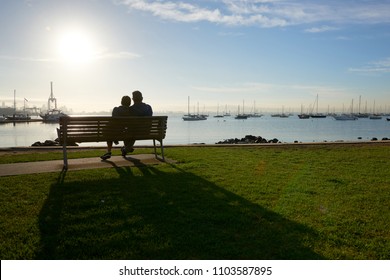 A Silhouette Of A Couple Sitting On A Bench By The Waters Edge Enjoying The Sun In Their Face And Vista Of Water And Boats.