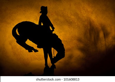 Silhouette Of Cowboy On A Bucking Bronco. Orange Yellow Sunset Background With Shadows Of Prairie Grass.