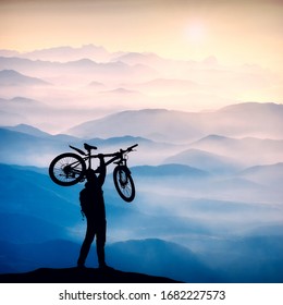 Silhouette Of Cyclist Who Hold Bicycle With One Hand Above The Head Against Sunrise In A Mountain Valley. Extreme Sport And Biking Concept.