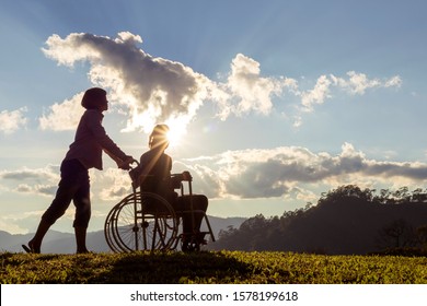 Silhouette Of Disabled Mother In Wheelchair And Daughter In Spring Nature At Sunset.