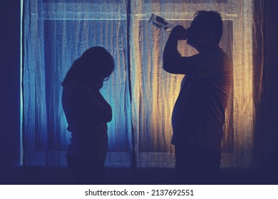 Silhouette Of A Drinking Man And A Sad Woman At The Night Window. Unhappy Couple Husband And Wife Having Drinking Problems In The Evening Light Of The Home Living Room