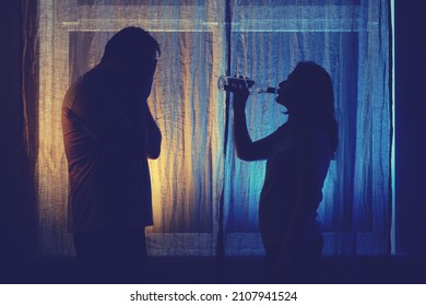 Silhouette Of A Drinking Woman And A Sad Man At The Night Window. Unhappy Couple Husband And Wife Having Drinking Problems In The Evening Light Of The Home Living Room