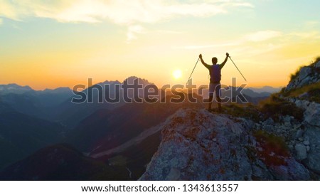 SILHOUETTE, DRONE, LENS FLARE: Golden morning sun rays shine on the ecstatic man hiking in the picturesque Slovenian Alps. Young male hiker celebrates reaching the mountaintop to see the sunrise.