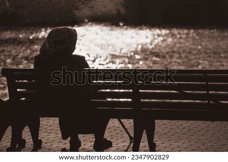 Silhouette of an elderly woman holding cane sitting on bench. Sunset on waterfront promenade. Concept old age and loneliness