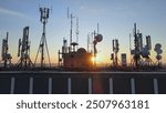 Silhouette of electromagnetic towers with satellite dish, microwaves and panel antennae at rooftop station during golden hour