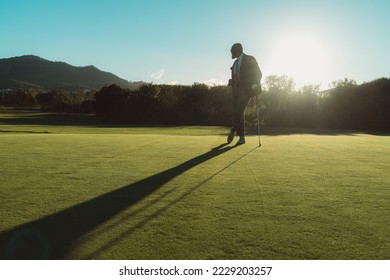 A silhouette of an elegant bald bearded black man in a tailored suit leaning on his gold club while standing on an empty lawn of a golf field, backlit by evening sun casting a long shadow on the grass - Powered by Shutterstock