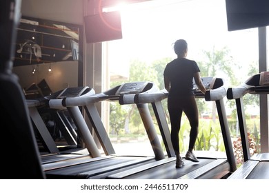 silhouette of excited woman exercising on treadmill machine, back view of asian woman running on treadmill machine in gym room with nature view