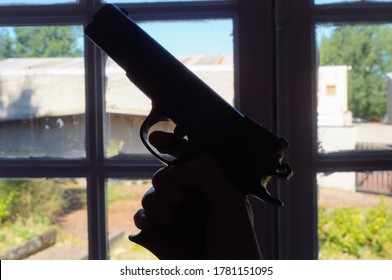 Silhouette Of A Fake But Realistic Gun (children's Toy Pistol) Featuring A Black Plastic Body Held By A Male Hand With The Finger On The Trigger And Pointed Upwards In Front Of A Multi-paned Window 