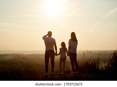 Silhouette Of A Family - Father, Mother And Daughter Standing On The Hill With Their Backs To The Camera And Looking To The Horizon On The Sunset