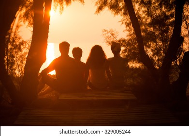 Silhouette Of A Family Sitting Down And Watching The Sun Setting Over The Sea. People Relaxing At The Seaside During Holiday And Enjoying A Beautiful Sunset Together.