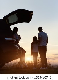 Silhouette Of The Family Of Three Enjoying The Sunset On The Top Of Hill Outside The City, Woman Is Sitting In The Car Trunk, Holding Jack Russell Terrier, Man Is Standing Embracing His Daughter