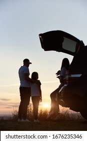 Silhouette Of The Family Of Three Enjoying The Sunset On The Hill Outside The City, Woman Is Sitting In The Car Trunk, Father Is Standing Embracing His Daughter