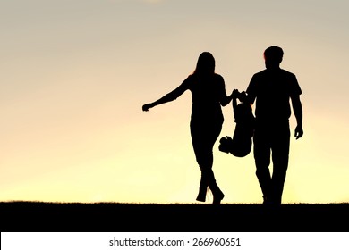 A Silhouette Of A Family Of Three People, Including Mother, Father, And Young Child Are Playing Around While Walking Outside At Sunset.