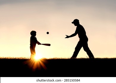 A silhouette of a father and his young child playing baseball outside, isolated against the sunsetting sky on a summer day.