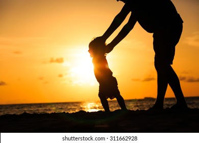 Silhouette of father and little daughter learning to walk at sunset beach