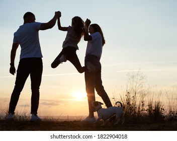 Silhouette of father, mother and their daughter spending fun day outside the city on the hill on the sunset, man and woman holding girl by hands and she is jumping, back view