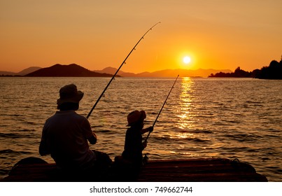 Silhouette Of Father And Son Or Boy And Man, Fishing On Wooden Pier At Sunset