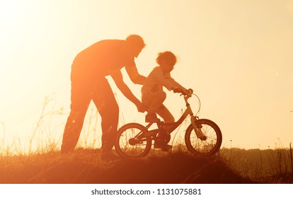 Silhouette Of Father Teaching Little Daughter To Ride A Bike At Meadow During Sunset. Caring Father Teaches Her Daughter To Ride Bicycle