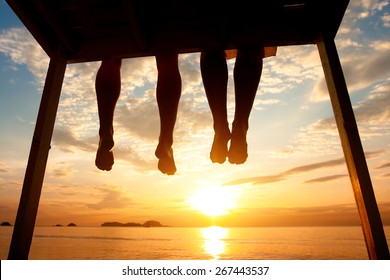 Silhouette Of Feet Of Couple Sitting On The Pier At Sunset Beach, Low Angle View