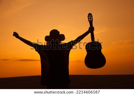 Silhouette of female musician holding guitar against sunset sky, summer nature festival