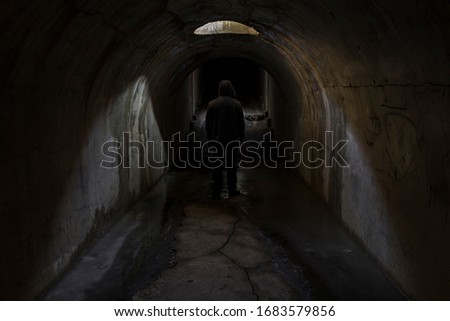The silhouette of the figure of a man going into unknown darkness in a gloomy dark old underground passage lit by sunlight through a hatch in the ceiling.