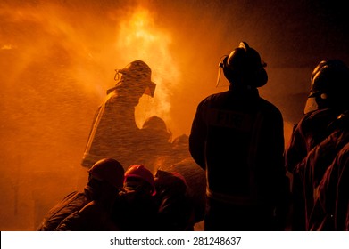 Silhouette Of Firemen Fighting A Raging Fire With Huge Flames Of Burning Timber
