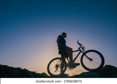 Silhouette Of A Fit Male Mountain Biker Riding His Bike Uphill On Rocky Harsh Terrain On A Sunset.