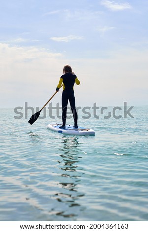 Silhouette fit sportsman stand up paddling on sup board or surfboard, enjoy to play extreme sport on holidays at sunlight beach. Copy space. Man in wetsuit holding paddles, concentrated, keep balance