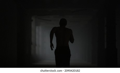 Silhouette Of Fitness Man Running In Dark Corridor. Male Runner Training In Loft Building. Muscular Athlete Doing Cardio Workout Indoors. Back View Athletic Guy Practicing Physical Exercises