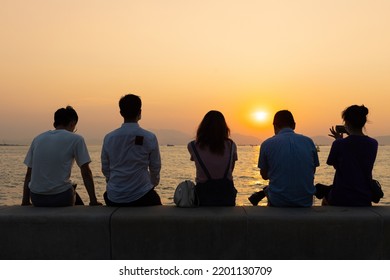 Silhouette Of Four Youngsters Students Boy And Girl Chat And Have Fun And Take Photos During Sunset In West Kowloon Waterfront Promenade, Hong Kong In Evening. Back Light Shot