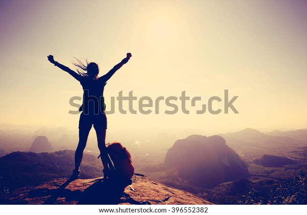 Silhouette of free cheering woman hiker open arms at\
mountain top cliff\
edge