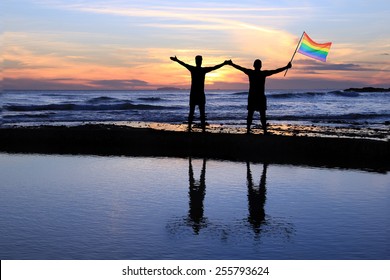 Silhouette Of A Gay Couple Holding A Rainbow Pride Flag At Sunset. 